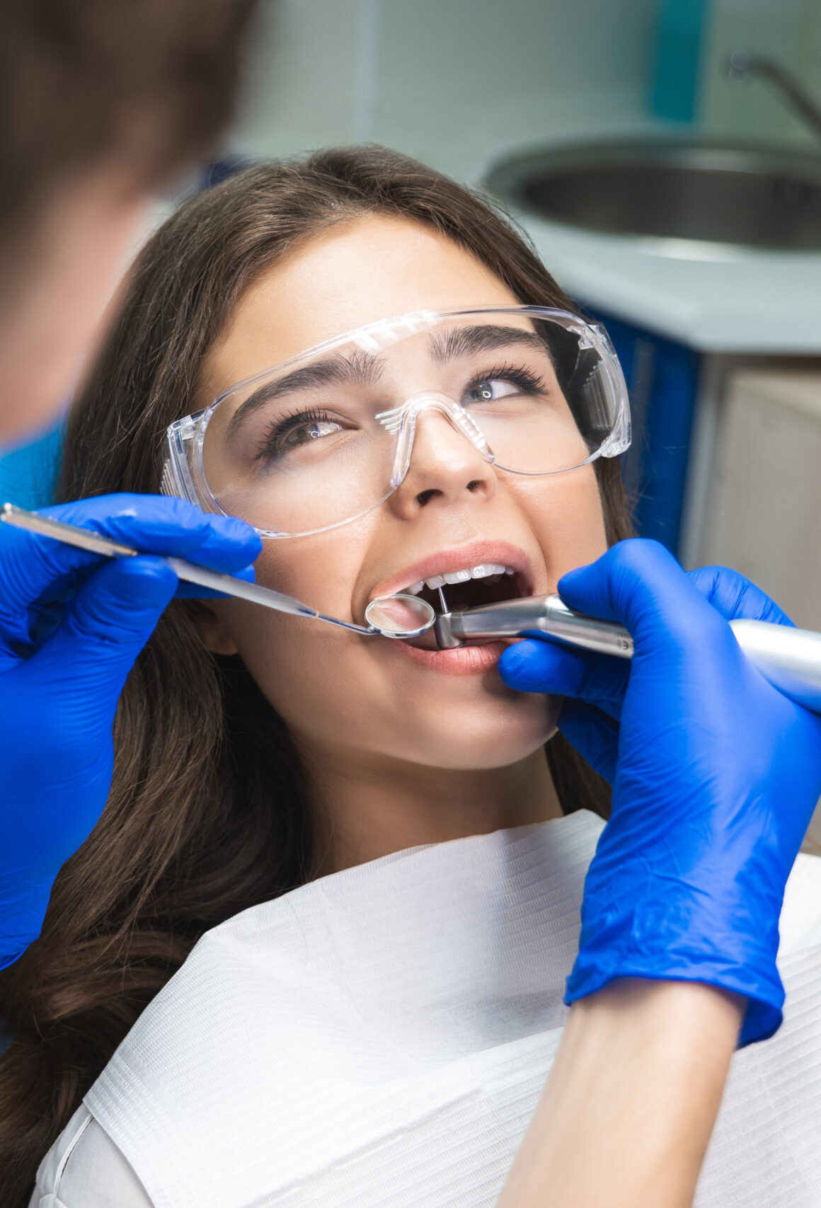 dentist in mask filling the patient's root canal while she is lying on dental chair in safety glasses under the medical lamp in clinic.