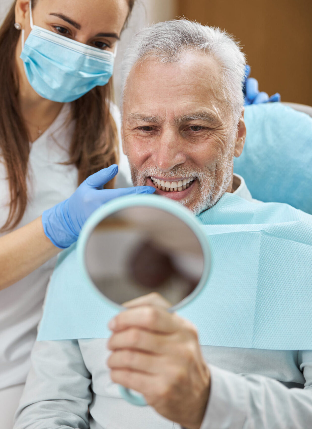 Elderly man and his young female dentist smiling while looking in the mirror in dental clinic