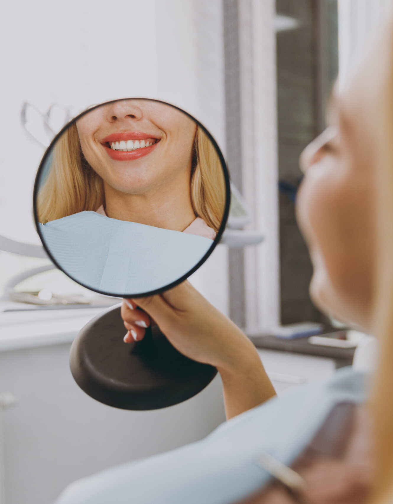 Young smiling satisfied happy woman 20s reflected in mirror sitting at dental office chair indoor light cabinet looking at teeth toothy smile whitening result blink Healthcare caries enamel treatment.