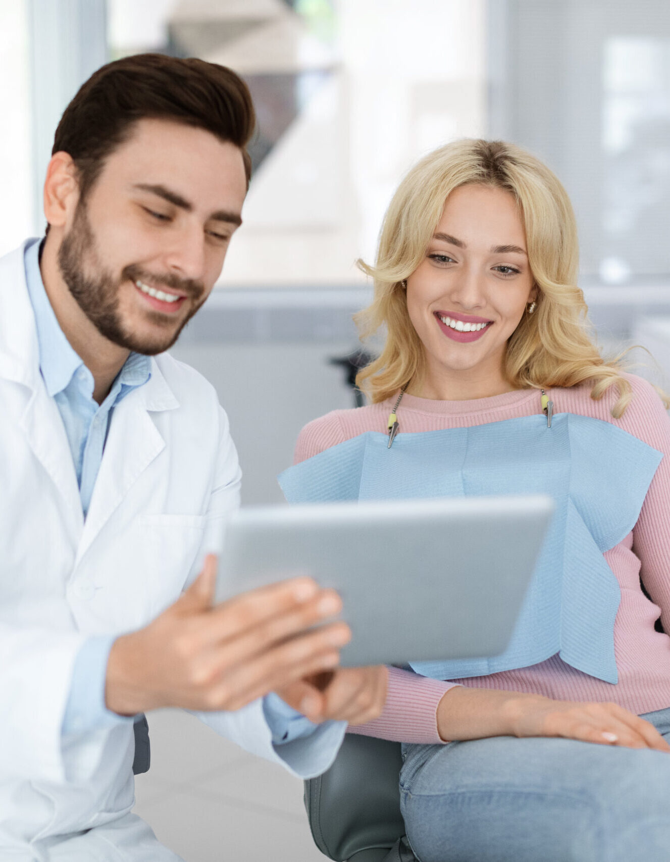 Cheerful young bearded man dentist discussing treatment method with beautiful blonde lady patient sitting at dental chair, looking at digital tablet. Modern technologies and healthcare concept